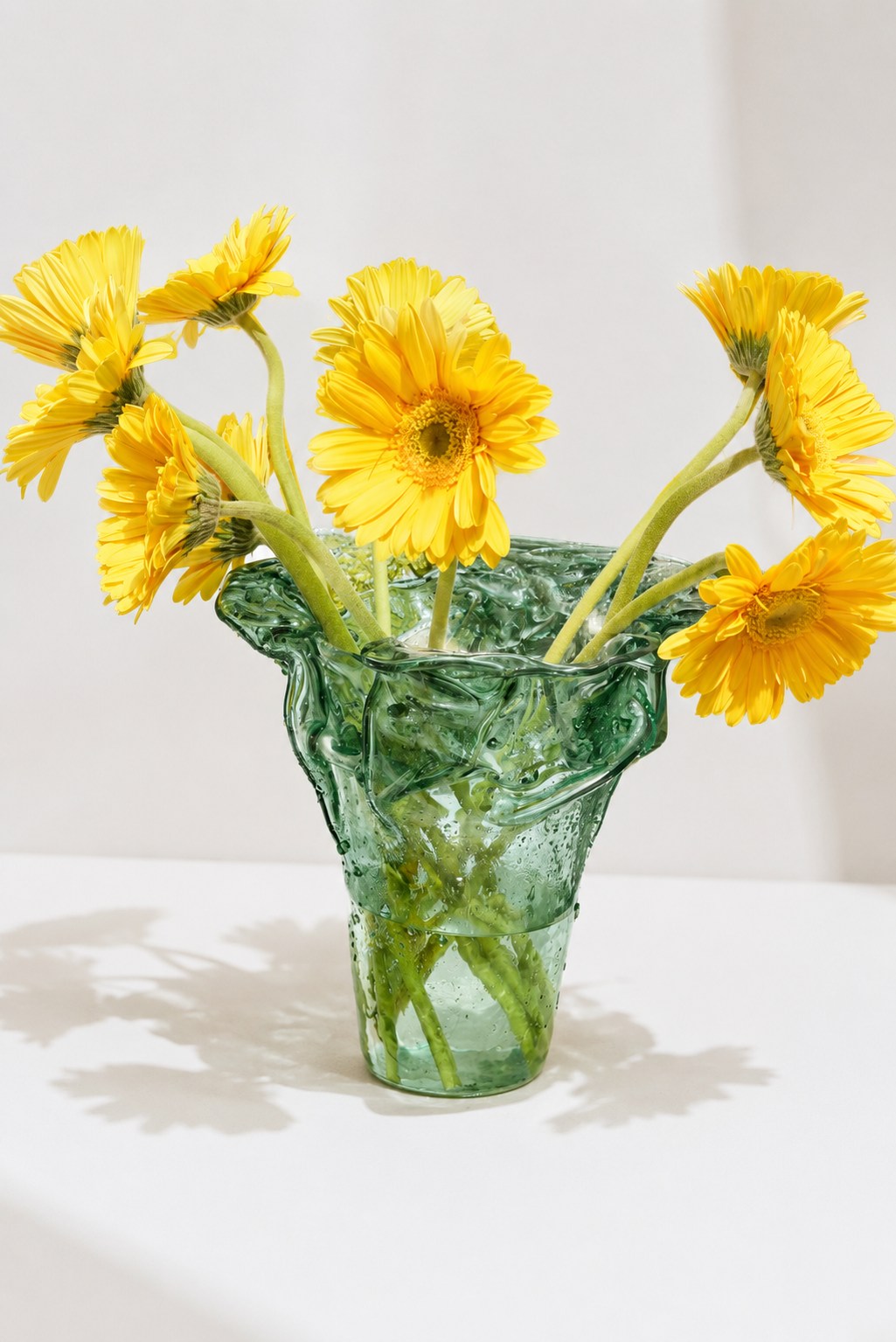 Florero Alto Irregular en vidrio verde con gerberas amarillas, sobre una mesa blanca con sombras de luz natural
